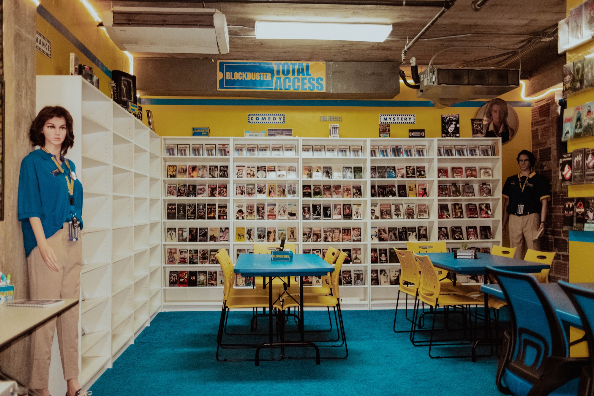 Inside the full-scale Blockbuster Museum set at Silver Sail Studios — yellow walls, VHS wall, and blue tables
